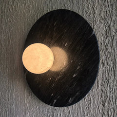 Halo wall-mounted light fixture featuring a large oval black alabaster stone base with a circular beige stone sconce, mounted on a textured gray wall.