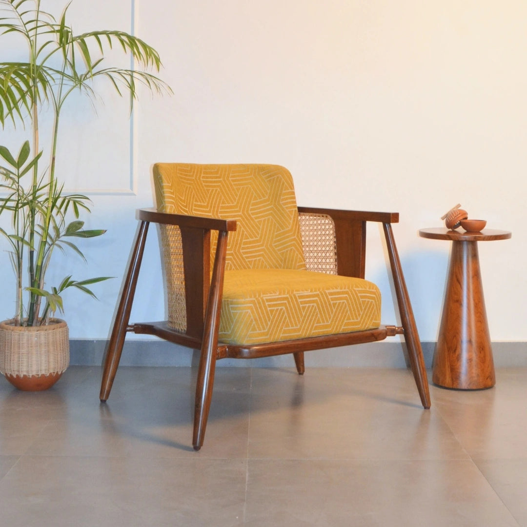 An accent chair with a vibrant yellow geometric pattern on the upholstery, teakwood frame, and cane details, positioned in a room with a plant and a wooden side table.