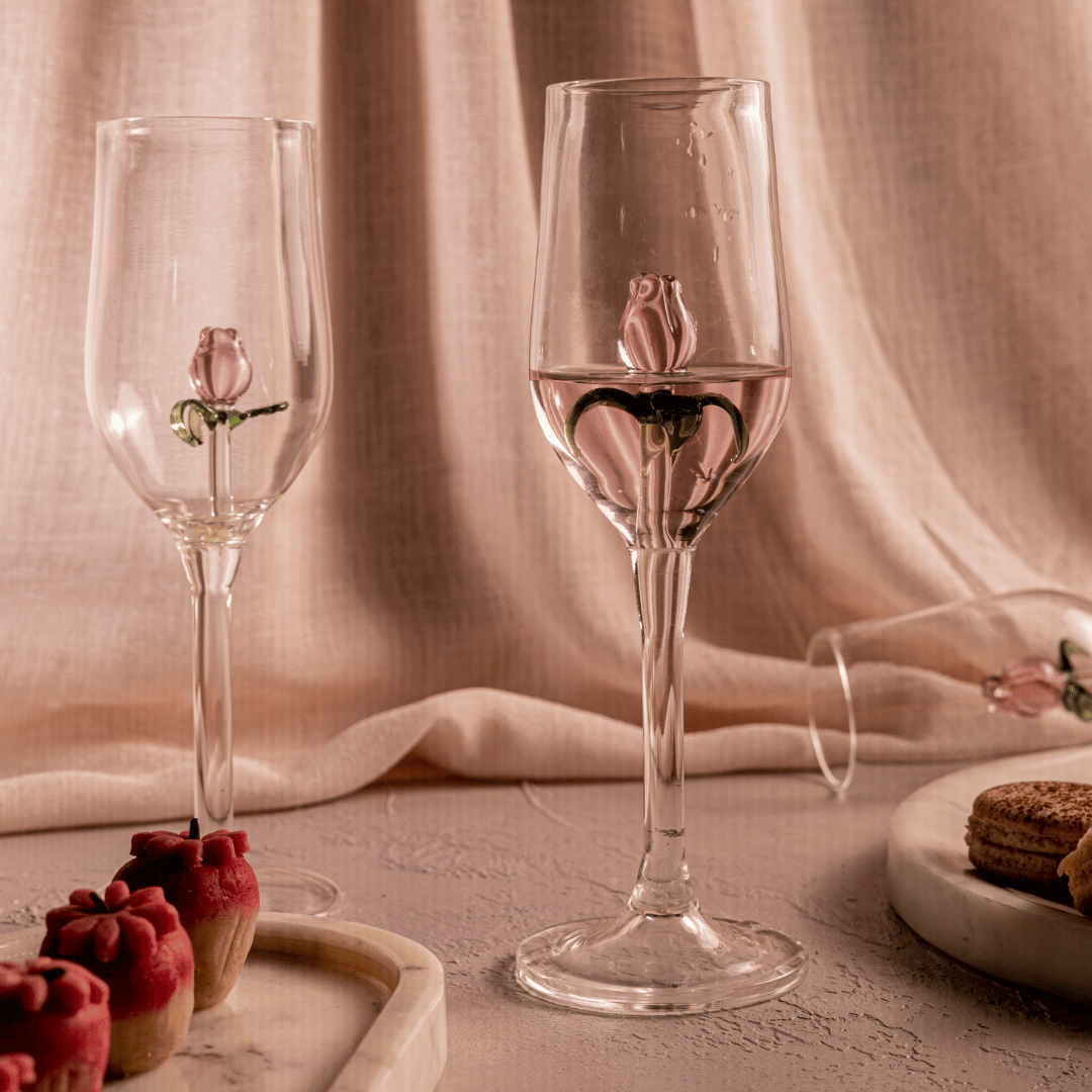 With Love = Two clear glass champagne flutes adorned with pink glass roses and green petals, placed on a table with a pink background, alongside a plate with red cupcakes.