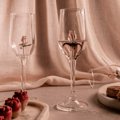 With Love = Two clear glass champagne flutes adorned with pink glass roses and green petals, placed on a table with a pink background, alongside a plate with red cupcakes.
