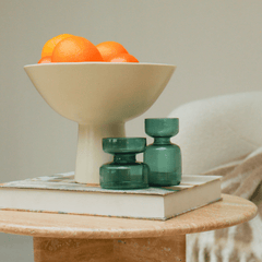 Two borosilicate glass mini vases (teal) in different shape and size, with a bowl of oranges in the background.