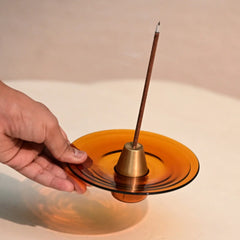 Hand placing an incense stick into a glass incense holder on a reflective surface.
