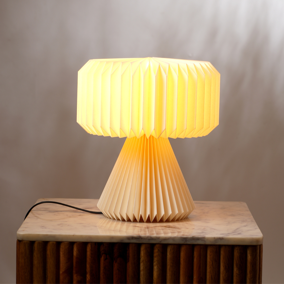 A beige origami paper table lamp with a conical pleated shade, illuminated, placed on a wooden surface.
