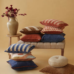 A styled collection of multiple decorative Holly Spice Velvet Stripe Cushion Covers in various patterns and colors, arranged on a woven bench and floor against a warm beige background. 