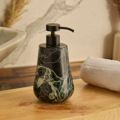A teardrop-shaped Spyder soap dispenser with a black metal pump and a dark green and black marble body, sitting on a wooden bathroom counter next to a white towel.