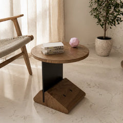 A round wooden side table with a black metal pedestal base and a wedge-shaped wooden foot, styled in a bright, modern room next to a woven chair and a potted plant.