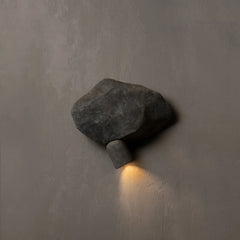A modern wall-mounted sconce shaped like a rough dark gray stone, casting a warm yellow light downward against a textured gray plaster wall.
