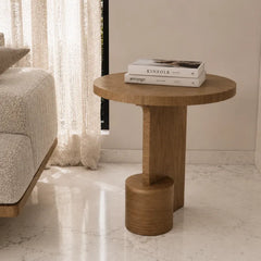 A round wooden end table with a sculptural pedestal base, styled in a bright, modern living room next to a textured sofa with books on top.