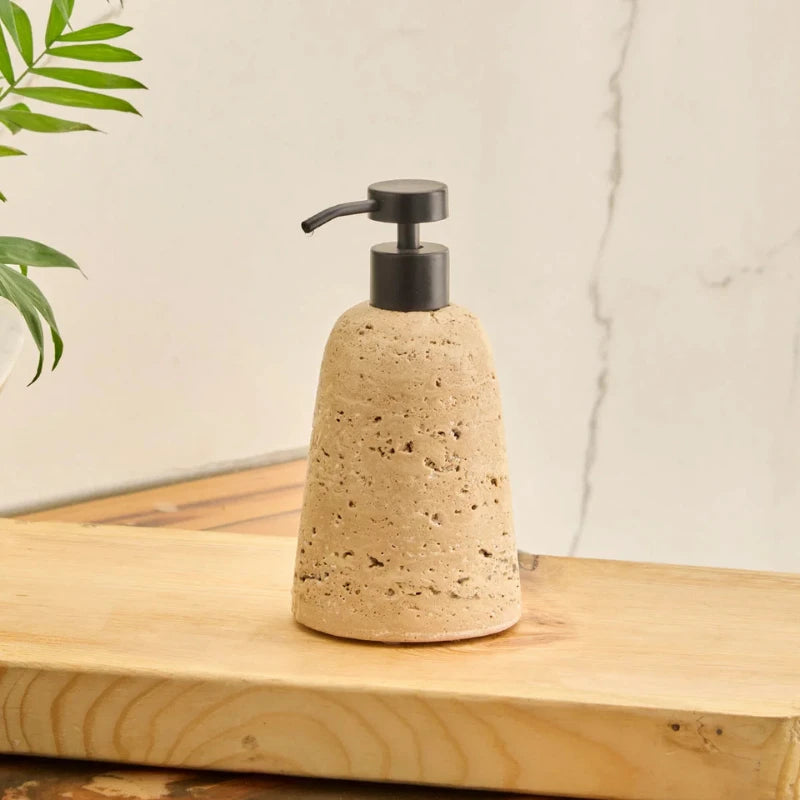 A natural travertine Eara soap dispenser with a black pump, sitting on a wooden board with a leafy plant in the background.