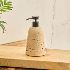 A natural travertine Eara soap dispenser with a black pump, sitting on a wooden board with a leafy plant in the background.