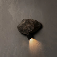 A rustic outdoor wall sconce featuring a dark textured stone top and a matte black metal base, casting a warm light downward against a textured gray concrete wall.