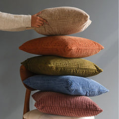 Five ribbed corduroy throw Luna Burnt Orange Velvet Cushion Covers in beige, terracotta, olive green, blue, and dusty rose are stacked on a wooden chair against a gray background, with a hand and arm holding the top beige pillow.