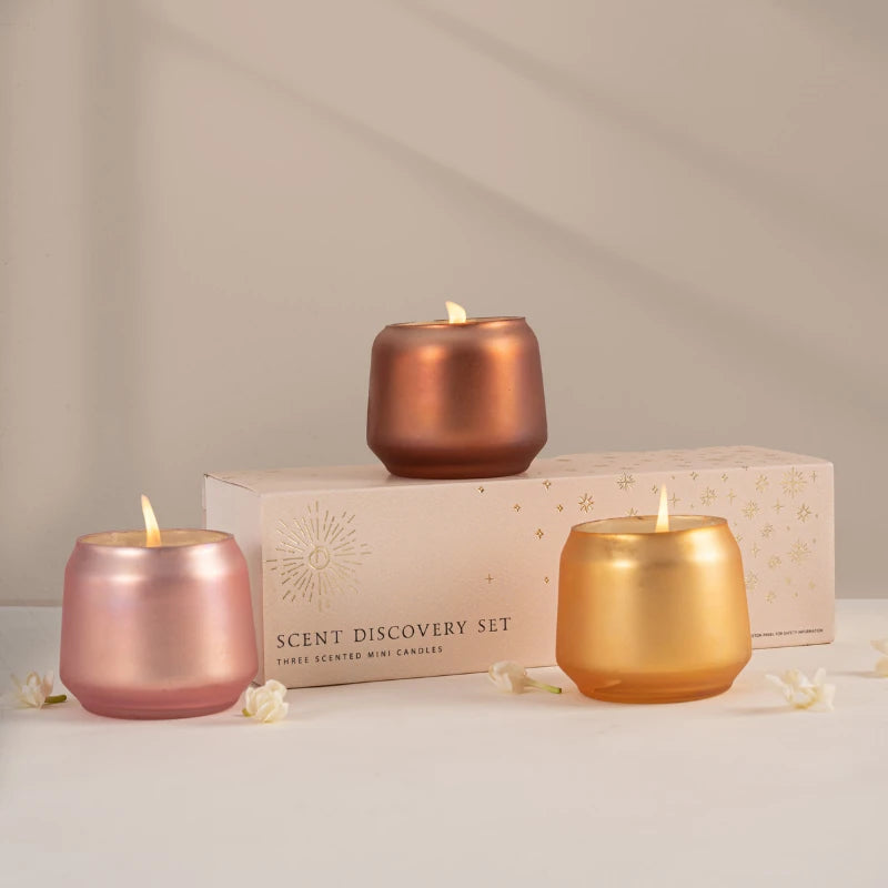 Three lit mini scented Votive Candle set in metallic rose gold, gold, and copper finish in Opulent Oud fragrance are arranged in front of their cream-colored gift box.