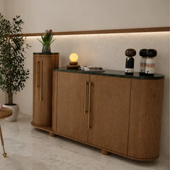 A modern wooden sideboard cabinet with a curved front, dark green marble top, and slim brass handles, styled in a minimalist interior with a potted plant, a small lamp, and decorative figures.