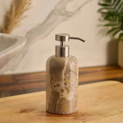 A marble-textured Koru Beige soap dispenser with a brushed silver pump, standing on a wooden surface in a bathroom setting.