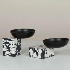 Two matte black ceramic Metropia Bowls displayed on top of black-and-white marbled cube pedestals against a neutral beige background.