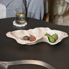 A wavy, two-compartment ceramic serving dish in a light beige stoneware finish, styled on a dark table