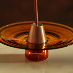 Copper incense holder with a wick on an orange glass base against a dark background