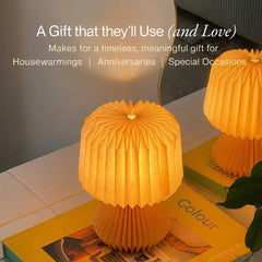 Decorative 2 warm glowing pleated Orilamp table lamps shaped like a small mushroom, shown on top of books in a styled indoor setting.