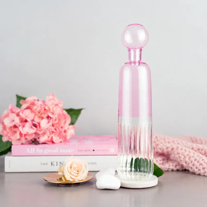 Firdaus Round Bottle in pink and clear glass with a ribbed lower half and a spherical stopper, styled on a tabletop with pink flowers, a book.