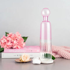 Firdaus Round Bottle in pink and clear glass with a ribbed lower half and a spherical stopper, styled on a tabletop with pink flowers, a book.