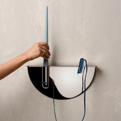 A hand holds a tall light blue taper candle in a modern semi-circular wall-mounted candle holder with a black and white design. The holder has a visible power cord and a blue plug, mounted on wall.