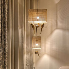 A pendant lamp with a handcrafted wooden bird design, featuring a two-tiered cane and teakwood structure, with lights inside. The lamp is hung against a backdrop of curtains with a light filter, creating a warm, ambient light.