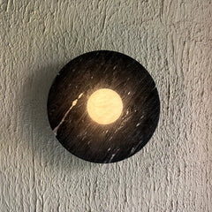 A round black halo alabaster wall sconce with a glowing warm yellow moon-like center, mounted on a textured off-white wall.