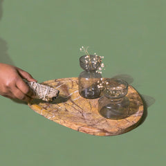 Hand holding a sage stick over a stone tray with glass containers on a green background