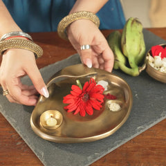 Taral Puja Thaali being arranged with red gerbera and lit diya on slate, antique brass finish, angled view