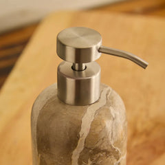 A close-up of a Koru Beige soap dispenser with a brushed stainless steel pump and a textured stone bottle with beige and gray marbling, set on a wooden surface.