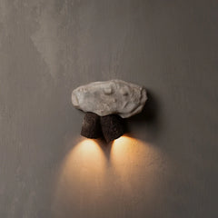 A rustic wall-mounted sconce featuring a textured light gray stone top and two dark brown cylindrical supports, casting a warm yellow light downward against wall
