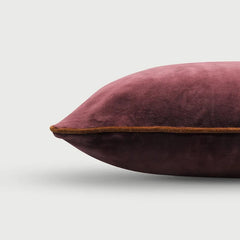 A close-up of a plush velvet throw pillow in a dusty rose or mauve color, featuring a contrasting tan leather piping detail along the edge, set against a clean light gray background.