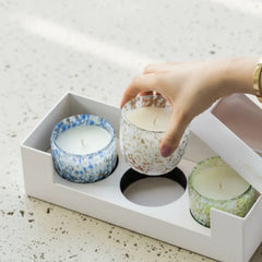 A hand is placing a glass jar candle into a white gift box alongside two other patterned candles. Votive candles are in blue, green, amber color with white speckles, on a speckled light countertop. 