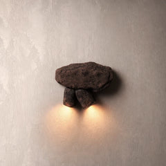 A rustic wall-mounted sconce shaped like a small stone animal, with a dark brown rock-like body and two smaller stones beneath it, casting a warm yellow light onto a textured light gray wall.