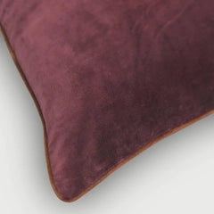 A close-up of a plush velvet throw pillow in a deep plum purple color, showing its soft texture and a thin brown piping edge against a light gray background.