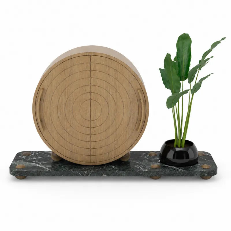 A round light wood decorative object with concentric circular grooves, displayed on a dark green marble base next to a small black planter with green leaves, on a white background.