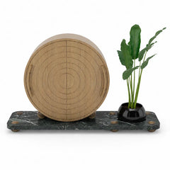 A round light wood decorative object with concentric circular grooves, displayed on a dark green marble base next to a small black planter with green leaves, on a white background.