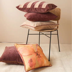 A stack of decorative throw pillows in various colors and textures, including a striped pillow, a ribbed burgundy pillow, a beige pillow, and a floral yellow pillow, arranged on and around a wicker chair with a black metal frame.