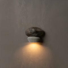 A wall-mounted sconce with a rough dark stone on top of a ribbed white ceramic base, casting a warm yellow light against a textured gray wall.
