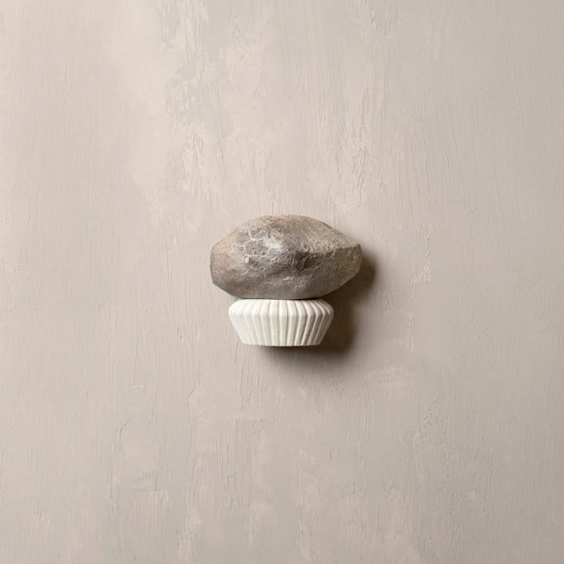 A wall-mounted decorative hook shaped like a small gray stone resting on a white ribbed cupcake liner, set against a textured light beige wall.
