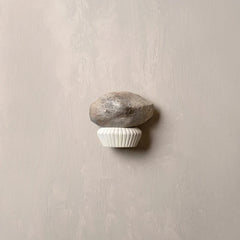 A wall-mounted decorative hook shaped like a small gray stone resting on a white ribbed cupcake liner, set against a textured light beige wall.