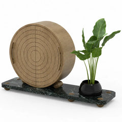 A decorative tabletop arrangement featuring a large round wooden cylinder with concentric target-style rings, resting on a dark green marble base next to a small black planter with green leaves, shown on a white background.