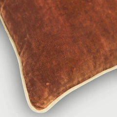 A close-up of a brown velvet throw pillow with a cream-colored piped edge, shown on a light gray background.