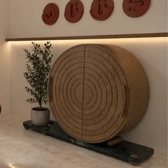 A large round wooden cabinet with concentric circle grooves on its front, displayed on a dark stone base in a styled interior setting with a potted plant and wall decor.