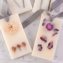 Close view of Two Wax Tablet, with dried flowers and rose petals and gray ribbon hangers, displayed on a white marble surface.
