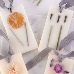 Flower Blossom & Lemongrass · Wax Tablet · Set of 4, with gray ribbon hangers, arranged on a light marble surface. Each features a small natural botanical element: a dried orange slice, three small cloves, a pale flower, and a purple flower.