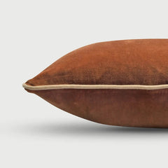 A close-up of a brown velvet throw pillow with a contrasting beige piping edge, shown against a light gray background.