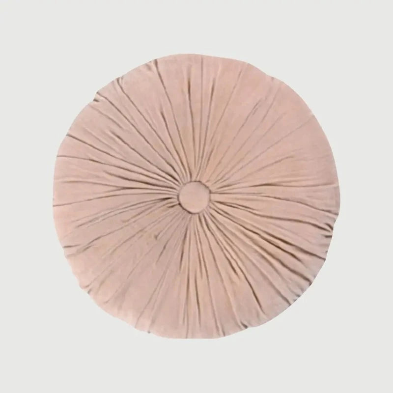 A round, light pink plush cushion with a central button and radiating pleated details, shown against a solid light gray background.