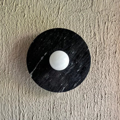 A modern circular halo alabaster wall sconce featuring a black marble outer ring with a white marble center, mounted on a textured beige plaster wall.
